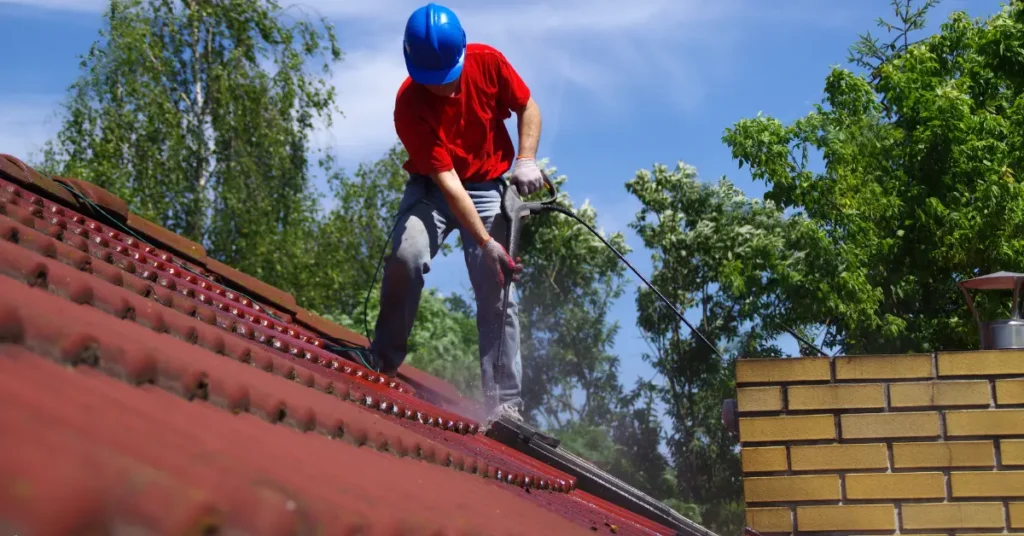 Outside window cleaning by a worker power washing a tiled roof under sunny skies.