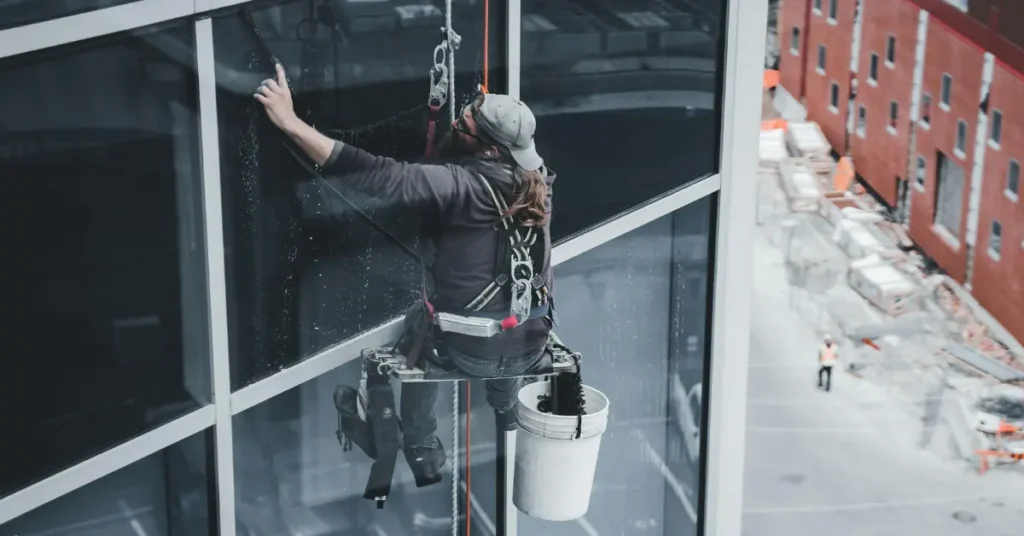 Window washing technician suspended on a harness scrubbing high-rise glass windows with a bucket and cleaning tools.
